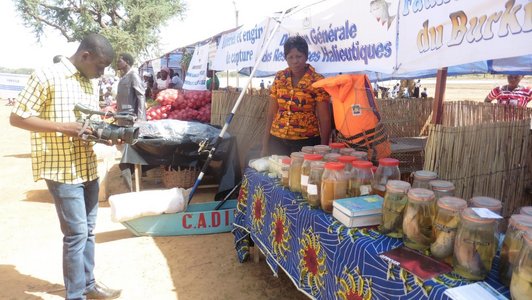 Man filming a market stall where a woman stands nearby