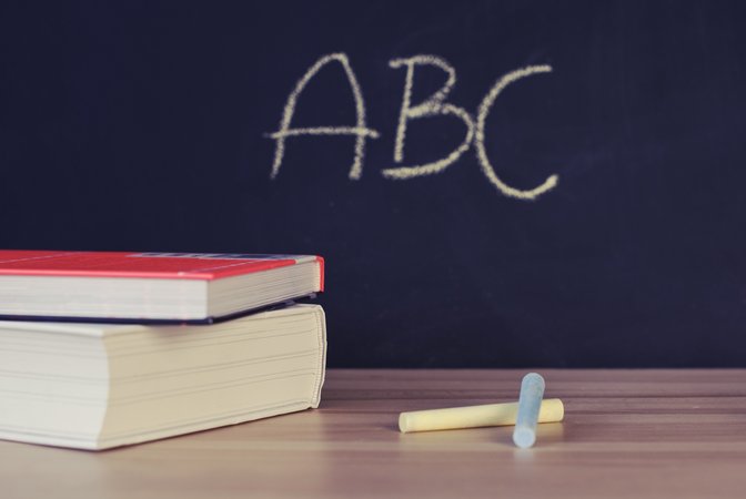 Tisch auf dem zwei Bücher und Tafelkreide liegen. Dahinter ist eine Tafel auf der ABC geschrieben ist.