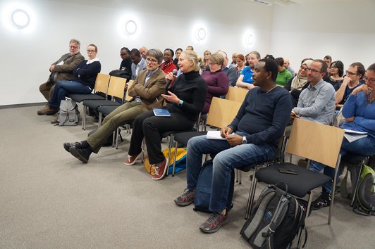 An audience sitting on chairs. One person is speaking into a microphone.