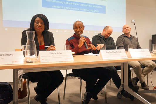 People sitting at a desk with name plates and glasses of water on it, in front of a large screen. One person is holding a microphone, apparently speaking to the audience. 