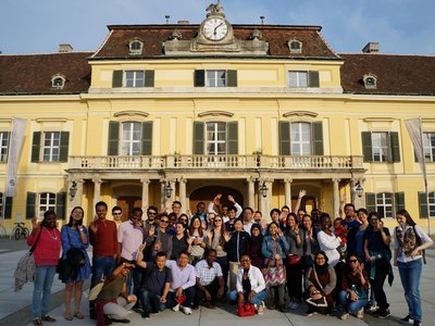 Many students from all around the world standing in front of yellowish old building in Laxenburg and waving to camer