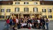 Many students from all around the world standing in front of yellowish old building in Laxenburg and waving to camer