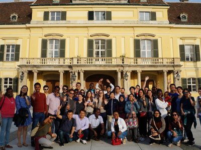 Many students from all around the world standing in front of yellowish old building in Laxenburg and waving to camer