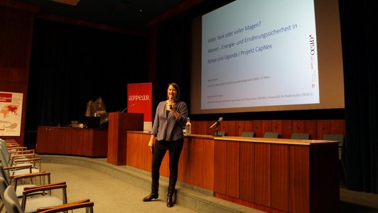 Doris Obrecht is standing in the front part of an auditorium, a microphone in her hand, speaking to the audience. A presentation is projected onto the large screen behind her, its title reads "Voller Tank oder voller Magen? Wasser-, Energie- und Ernährungssicherheit in Kenya und Uganda. Projekt CapNex".