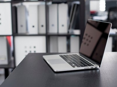 An open laptop is on a black table. In the background you can see a shelf with many different folders.