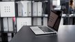 An open laptop is on a black table. In the background you can see a shelf with many different folders.