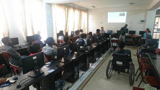 Group of people sitting and workplaces looking at a man who is standing in front of a presentation on the wall