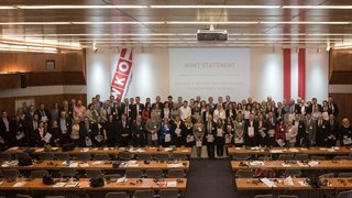 A large group of people take a group photo in a lecture hall. Most of them hold a piece of paper in their hand.