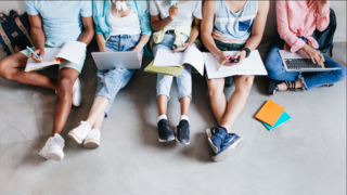 am Boden sitzenden und in Büchern arbeitende Jugendliche. Nur Unterkörper sind zu sehen. 