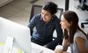 Multiracial colleagues indian and caucasian young women having coffee break sitting together at desk in office. Diverse students interns friends looks at pc screen talking discussing working moments