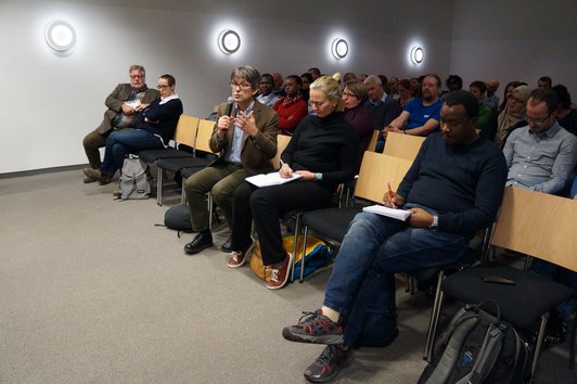 A sitting audience of approximately 30 people. One person is holding a microphone and giving a speech.