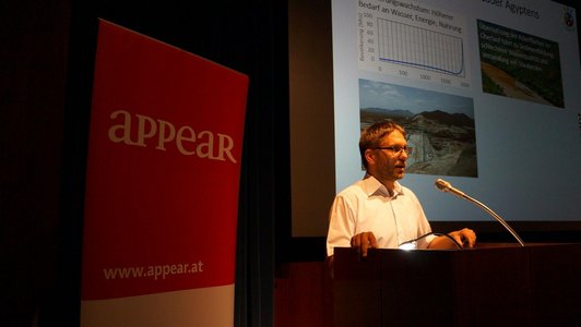 A man is giving a speech at a speaker's desk. There are a large screen showing a presentation and a roll-up announcing the "appear" programme behind him.