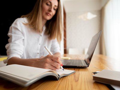 Person macht sich Notizen. Vor Ihr steht ein Laptop.