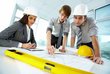 A woman and two men wearing construction site hats discuss a construction plan.