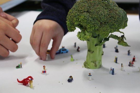 Kleine Figuren stehen auf einem Tisch, daneben Brokkoli, der größenverhältnismäßig wie ein Baum aussieht. Eine Hand ragt ins Bild hinein.