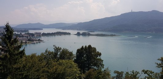 Der Wörtersee ist in der Mitte, im Hintergrund sind ein paar Berge auf einem ist der Pyramidenkogel zusehen. Im Vordergrund sind Bäume und Links sind einige Hotels und andere Gebäude am Seeufer zu sehen. 