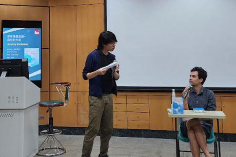 Eine stehende Person (links) und schaut nach rechts zu einer, an einem Tisch sitzenden Person, dahinter eine Leinwand