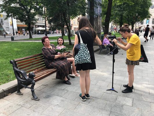 Two people on a park bench. A camerawoman with a tripod is filming them, another is talking to them.