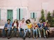 Seven students are sitting on a bench against the wall of a house.