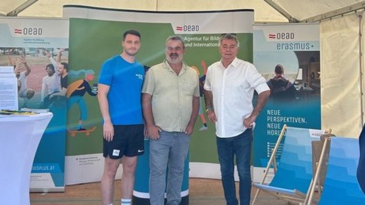 Gruppenbild mit drei Männern vor einem Messestand im Freien. Die Sonne scheint.