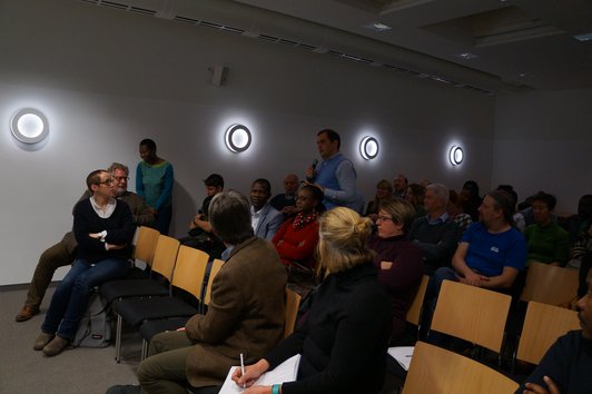 An audience sitting on chairs. One man is standing and holding a microphone, speaking to the others.