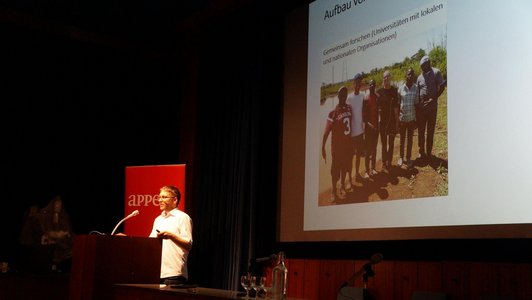 A man is giving a speech at a speaker's desk. A presentation is projected onto a large screen behind him.