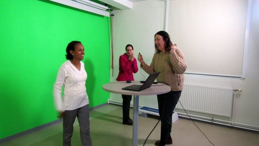 One person standing in front of a green wall, two other persons watching.