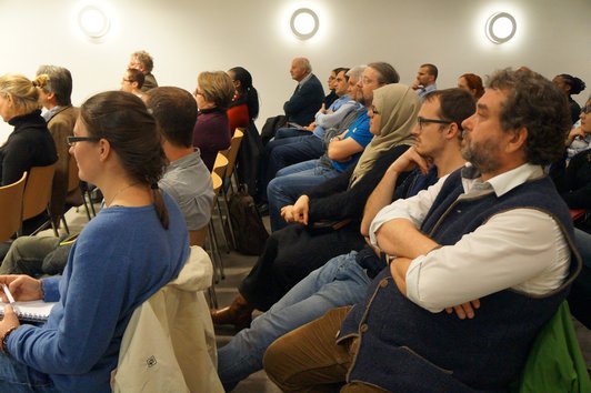 An audience sitting on chairs in a room with white walls and a carpeted floor.