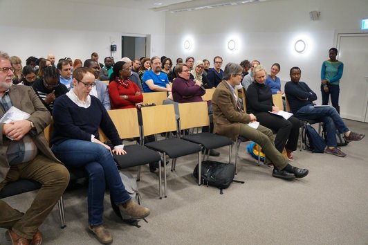 An audience sitting on chairs. One person is speaking into a microphone. 