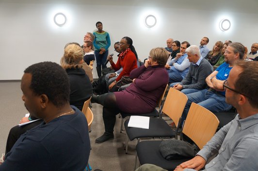 An audience sitting on chairs. One person is standing, one person is speaking into a microphone.