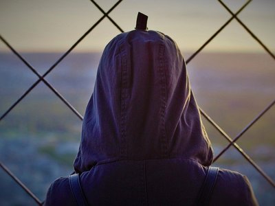 Kopf einer Person mit Schleier oder Kapuze von hinten, die vor einem Zaun steht.