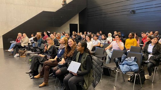 Das Publikum eines Vortrages sitzt in schwarzen Stühlen in einem großen Hörsaal. Es sind ca. 40 Personen zu sehen