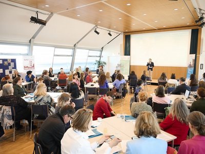 Zahlreiche Personen an Tischen, Moderator vorne auf der Bühne