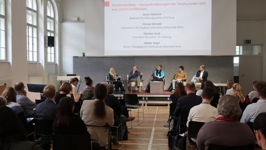 Eine kleine Bühne in einem Saal auf der fünf Personen in Sesseln sitzen. Einer von ihnen hat ein Mikrofon in der Hand. Auf der Wand hinter ihnen ist eine Leinwand mit einer PowerPoint darauf (Herausforderungen beim Einstig in Hochschulen). Im Vordergrund ist ein voller Saal mit Zuschauern zu erkennen.