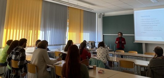 Vortragende vorne an Tafel mit Präsentation, Zuhörende in Tischreihen