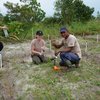 A woman and a man are on their knees on a field. The woman has a plastic bag with some earth in her hand and the man has a device in his hand. Next to them stands another man, who also has a plastic bag in his hand.