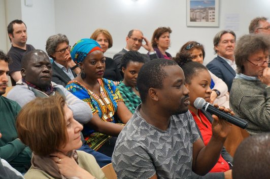 The picture shows a sitting audience. One person is speaking into a microphone.