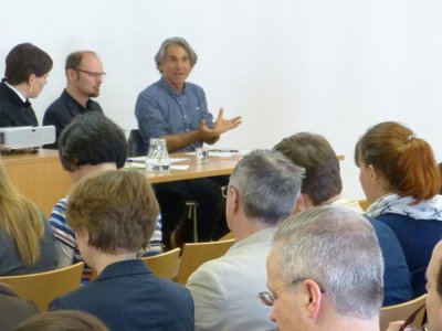 Auftaktveranstaltung: Dialog hochschulischer Lehre. Viele Personen sitzen in einem Saal und schauen zum Podium, auf dem drei Personen sitzen, eine davon spricht gerade