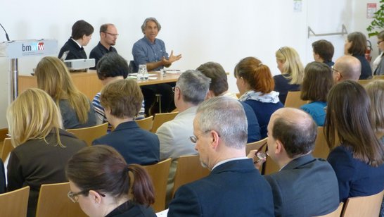 Auftaktveranstaltung: Dialog hochschulischer Lehre. Viele Personen sitzen in einem Saal und schauen zum Podium, auf dem drei Personen sitzen, eine davon spricht gerade