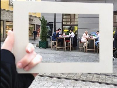 Someone holds a picture frame in front of the camera. Through the picture frame you can see a table with three businessmen sitting outside a restaurant and talking