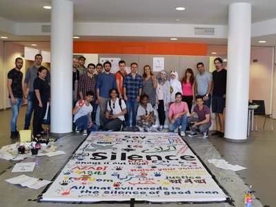  A group of people stand in front of a work of art about silence. In the form of a large fabric square which has been described in several languages.