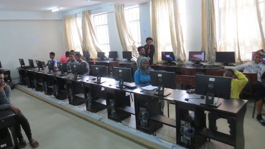 Wokrplaces with computers standing in rows and some people sitting at the tables