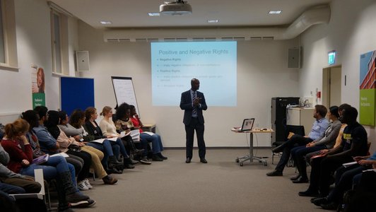 People sitting in chairs on the left and on the right, looking at a male presenter in the middle