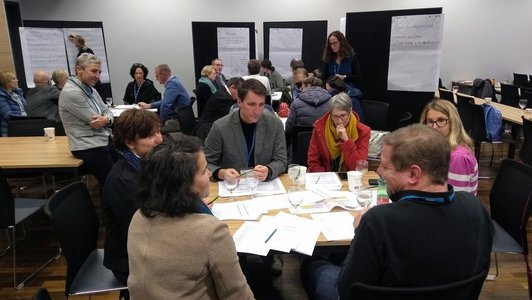 ein Raum mit vielen Personen, die an Tischinseln an Gruppenarbeiten arbeiten