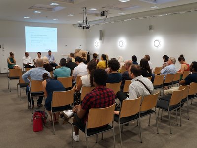 Saal mit Menschen, die auf Stühlen sitzen und einer Podiumsdiskussion lauschen