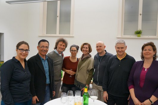 A group of eight people is standing around a table with glasses on it, smiling into the camera.