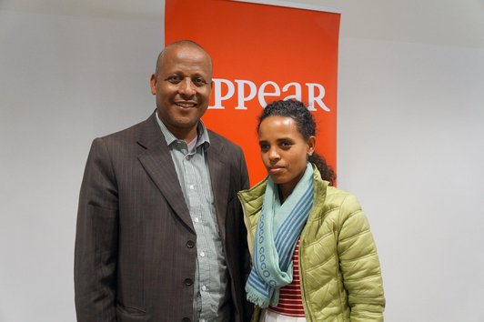 Two people are standing in front of a red-coloured roll-up reading "appear".