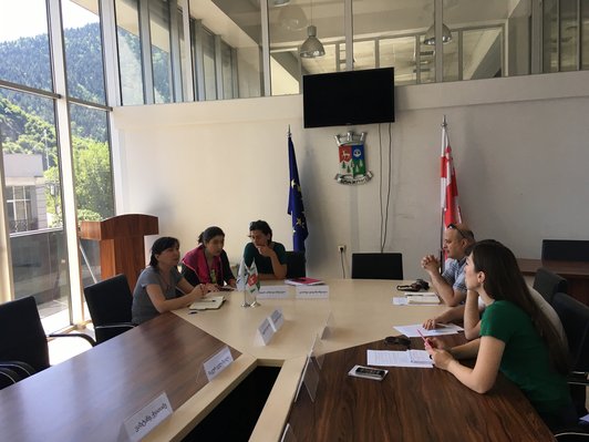 Group of people are sitting at a big table under a screen talking to each other
