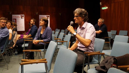 A man is sitting amidst an audience and speaking into a microphone. 