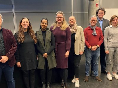 Group photo with nine people, four of them men and five women, in a lecture hall in front of the desk.